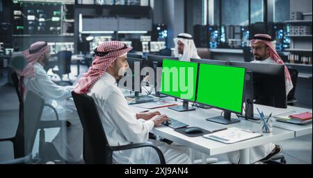 Group of Middle Eastern Muslim Men Working in Research Center, Collaborating on an Online Project. Young Arab Gulf Male in Traditional Clothes Using Computer with Green Screen Mock Up Display Stock Photo