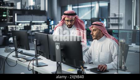 Young Team of Arab Specialists Working on Computers and Having a Conversation at a Workplace. Two Male Software Developers Discussing a Solution for Their Generative Artificial Intelligence Project Stock Photo
