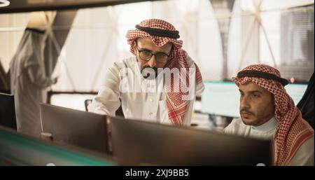 Arab Graphic Designer Working on Computer, Discussing Project with Team Leader. Middle Eastern Colleagues Working in Modern Office, Wearing Traditional White Thobes and Kaffiyeh Stock Photo
