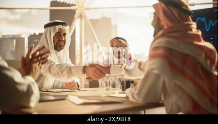 Middle Eastern Business Partners Striking a Successful Deal at a Corporate Modern Meeting Room. Two Arab Men Shaking Hands, Managers in Traditional White Robes Celebrating and Clapping Hands Stock Photo