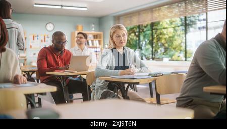 Grown Up Men and Women Getting Specialty Knowledge on an Evening Course in College. Adult Class Writing Down Notes in Notebook and Using Laptop Computers. Teacher Walking Between Desks Stock Photo