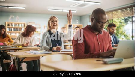 Diverse Mature Students Learning in Classroom, Using Laptops and Writing in Notebooks. Female Raising Hand and Asking Teacher a Question During a Lecture in Adult Education Course in School Stock Photo