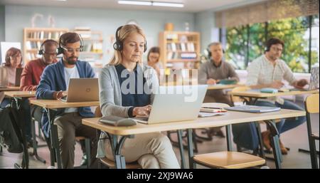 Diverse Mature Students Learning in Classroom, Sitting Behind Desks with Headphones, Using Laptops and Writing in Notebooks. Teacher Giving an Adult Education Course Remotely Online Stock Photo