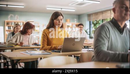 Adult Education Center: Young Beautiful Female Student Learning in Classroom, Using Laptop Computer to Write Down Notes. Group of People Taking a Workshop on Improving Professional Work Skills Stock Photo