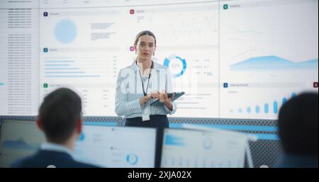 Female Project Manager With Tablet Computer Is Giving Presentation To Diverse Employees In Front of Big Digital Screen In Monitoring Room. Team Of Data Scientists Listening To New Objectives, Tasks. Stock Photo