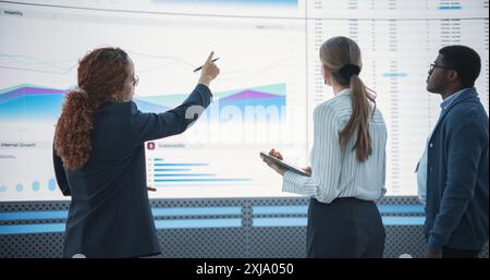 Diverse Team Of Data Scientists Analyzing Information And Graphs On Big Digital Screen In Modern Monitoring Office. Multiethnic Colleagues Discussing Financial Strategies, Risk Factors, and Deals. Stock Photo