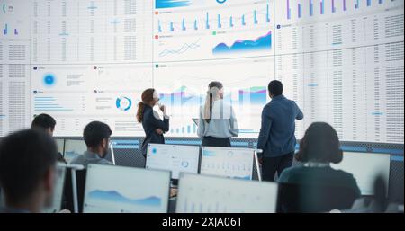 Diverse Team Of Data Scientists Having A Meeting And Discussing Reports In a Modern Monitoring Office With Analytics Feed On a Big Digital Screen. Big Data Company With Employees Behind Computers Stock Photo