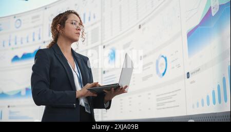Successful Hispanic Businesswoman Holding Laptop Computer And Reviewing Financial Reports On Digital Screen In Monitoring Office. Female Leader Running Deep Learning, Artificial Intelligence Company. Stock Photo