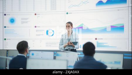 Caucasian Female Project Manager With Tablet Computer Is Giving Presentation To Diverse Employees In Front of Big Digital Screen In Monitoring Room. Team Of Data Scientists Listening To New Objectives Stock Photo