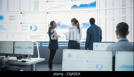 Diverse Team Of Financial Data Analysts Studying Information And Graphs On Big Digital Screen In Modern Monitoring Office. Multiethnic Employees Working Infront Of Desktop Computers In Consulting Firm Stock Photo