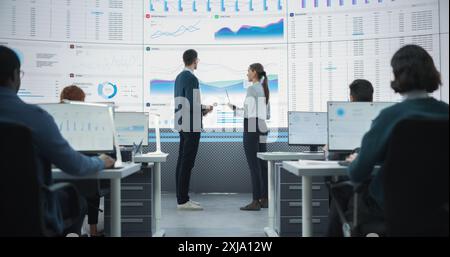 Zoom Out Back View Shot Of Male And Female Analysts Discussing Financial Data On Big Digital Screen In Monitoring Office. Diverse Employees Working Behind Desktop Computers In Venture Capital Company. Stock Photo