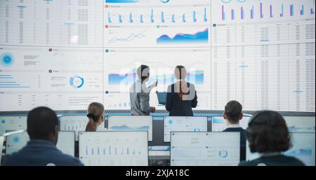 Wide Shot of Diverse Male And Female Data Analysts Discussing Graphs And Reports On Big Digital Screen In Monitoring Office. Multiethnic Employees Working On Desktop Computers for AI Company. Stock Photo