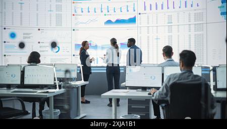 Diverse Team Of Data Scientists Analyzing Information And Graphs On Big Digital Screen In Modern Monitoring Office. Multiethnic Employees Working Infront Of Desktop Computers In Consulting Firm. Stock Photo