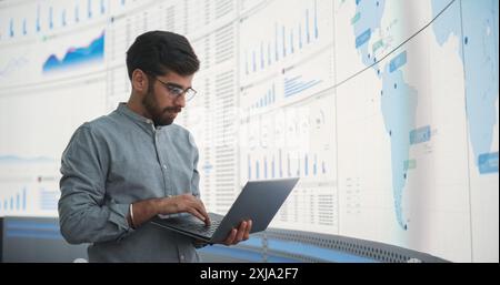 Indian Male Product Distribution Expert Holding Laptop Computer, Analyzing World Map On Big Digital Screen In Monitoring Office. Successful Man Looking For Efficient Routes For International Delivery. Stock Photo