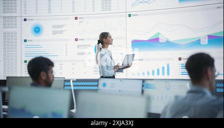 Female Data Scientist Using Laptop And Analyzing Information And Graphs On Big Digital Screen In Monitoring Office. Caucasian Woman Working For Big Data Company, Diverse Employees Behind Computers. Stock Photo