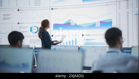Female Data Scientist Using Laptop And Analyzing Information Graphs On Big Digital Screen In Monitoring Office. Hispanic Woman Working For Big Data Company, Diverse Employees Behind Desktop Computers Stock Photo