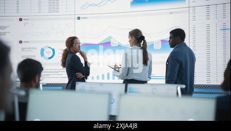 Diverse Team Of Data Scientists Having A Meeting And Discussing Reports In a Modern Monitoring Office With Analytics Feed On a Big Digital Screen. Deep Learning Company With Employees Behind Computers Stock Photo