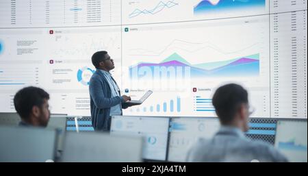 Male Financial Data Analyst Using Laptop And Studying Information And Graphs On Big Digital Screen In Monitoring Office. Black Man Working For Big Consulting Firm, Diverse Employees Behind Computers. Stock Photo