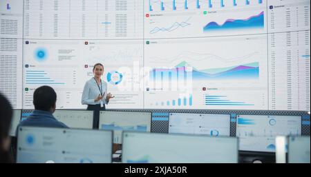 Caucasian Female Data Scientist With Tablet Computer Is Reviewing Reports Of Diverse Employees on Big Digital Screen In Monitoring Room. Team Of Analysts Listening To New Objectives And Tasks. Stock Photo