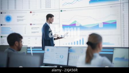 Male Senior Financial Analyst Reviewing Information And Reports on Big Digital Screen in Modern Monitoring Office. Diverse Employees Working Behind Desktop Computers In Investment Management Company. Stock Photo