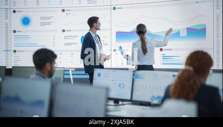 Female Data Scientist And Male Project Manager Discussing Reports And Graphs On Big Digital Screen In Monitoring Office. Diverse Employees Working Behind Computers in Artificial Intelligence Company. Stock Photo