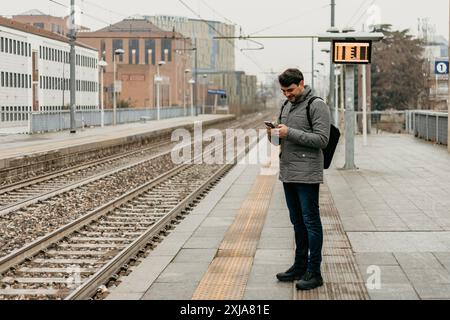 A man stands on a train station platform checking his phone while waiting for a train. Stock Photo