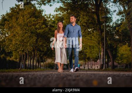 A couple walks hand in hand on a pathway through a park, the sun setting behind the trees. Stock Photo