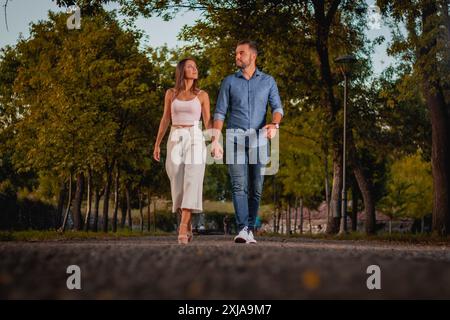 A young couple walks hand-in-hand down a path through a park, with trees lining either side. The sun is setting in the background. Stock Photo