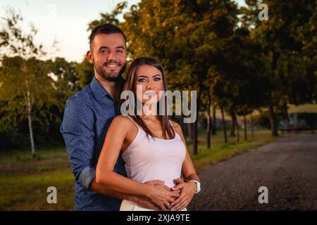 A young man and woman stand together in a park, with the man's arm around the woman's waist. They are both looking at the camera and smiling. Stock Photo