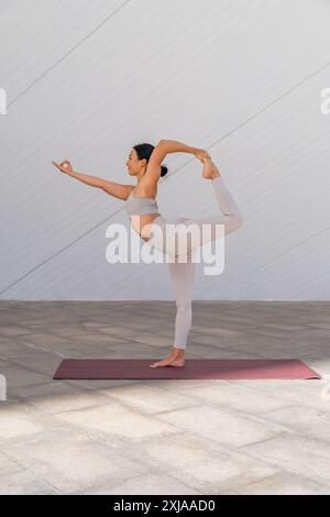 Brunette woman dressed in sportswear performing Natarajasana yoga pose or dancer's pose on modern, minimalist background. Stock Photo