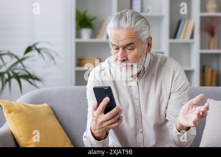 Elderly man sitting on couch using mobile phone, looking confused and expressing frustration with hand gesture. Home interior background with bookshelf and plants. Stock Photo