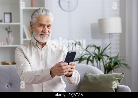 Portrait of an older gray-haired man in a white shirt sitting on the couch at home, holding a mobile phone and smiling at the camera. Stock Photo