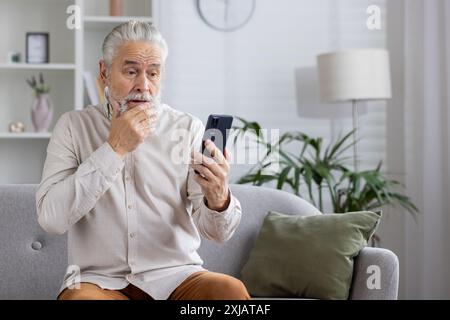 Worried elderly man holding smartphone and touching chin, sitting on sofa in living room. Concept of technology, communication, and senior's reactions. Stock Photo