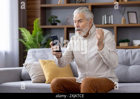 Shocked senior man sitting on sofa at home and looking surprised at phone screen, got bad news, spreads hands. Stock Photo