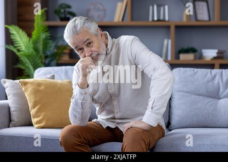 Portrait of a serious and pensive senior man sitting on the couch, leaning his head on his hand and looking sadly into the camera. Stock Photo