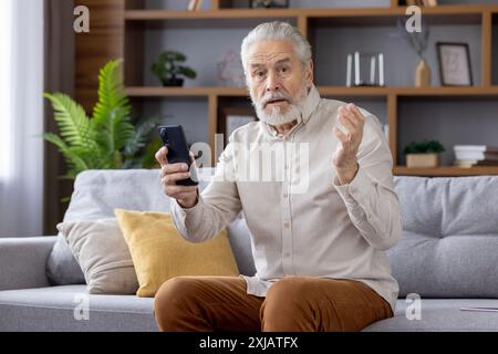 Portrait of a frustrated and disappointed sad man grandfather sitting at home on the sofa, holding the phone and looking sadly at the camera, spreading his hands. Stock Photo