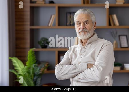 Portrait of a serious gray-haired old man with a beard standing in the office in a shirt, crossing his arms on his chest and confidently looking at the camera. Stock Photo