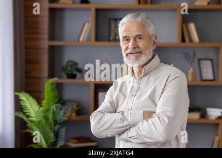 Portrait of a scary gray-haired man standing in a home office, crossing his arms on his chest and looking confidently and smiling at the camera. Stock Photo
