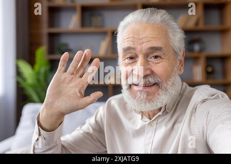 Close-up photo of an older gray-haired man with a beard talking on the phone camera, taking a selfie at home, greeting and waving. Stock Photo