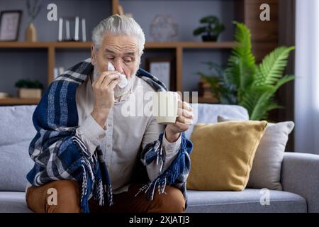 An elderly man is not feeling well, sitting at home on the sofa covered with a blanket, holding a cup with a drink in his hands and wiping his nose with a napkin from a runny nose. Stock Photo