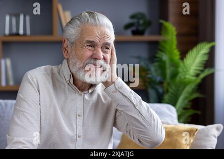 Close-up portrait of an older man suffering from severe ear pain, sitting on the sofa at home and grimacing, holding his hand behind his head. Stock Photo