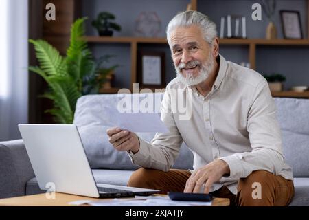 Portrait of smiling senior gray-haired man sitting on sofa at home and checking bills and documents, using laptop and calculator, looking at camera. Stock Photo