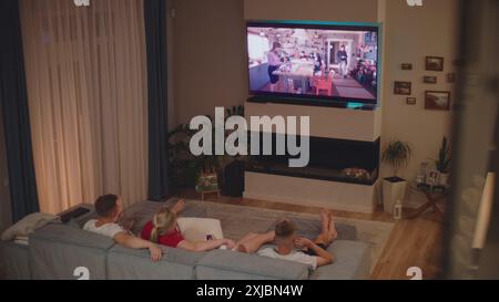 ZAPORIZHYA, UKRAINE - JUNE 7, 2024: High angle view of Caucasian family lying on sofa in cozy living room in the evening watching TV show or movie on Netflix. Man comes and lies down on sofa with his wife and children. Home theater. Stock Photo