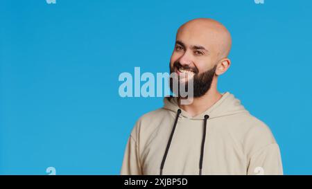 Middle eastern guy greeting people in front of camera, waving hello and smiling over blue background. Young person posing with confidence in studio, feeling confident and joyful. Camera 2. Stock Photo