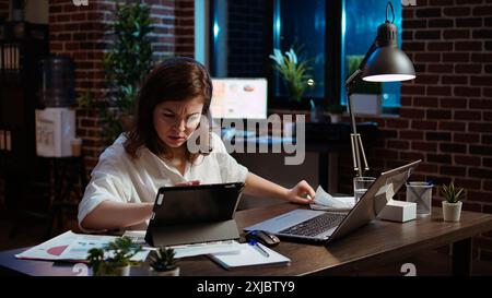 Businesswoman researching for company project, crosschecking between paperwork files, tablet and laptop screens. Employee looking over accounting figures on electronic devices displays in office Stock Photo