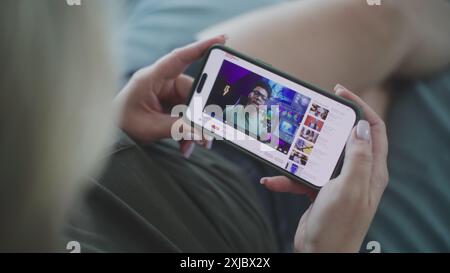 Close up of Caucasian woman watching video about color grading using mobile phone while lying on sofa. Video hosting website with video content about color correction displayed on smartphone screen. Stock Photo