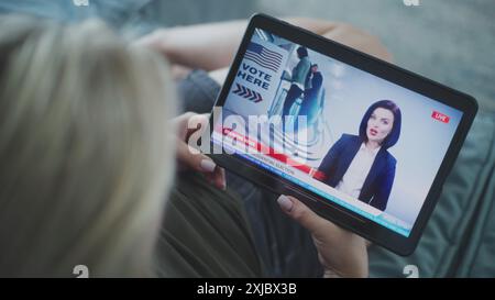 Tablet screen shows TV news broadcast with female newscaster. Caucasian woman watching breaking news about United States presidential elections using digital tablet while lying on sofa. Close Up. Stock Photo
