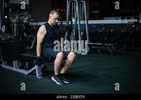A man in black workout attire lifts weights while sitting on a bench in a dimly lit gym. Stock Photo