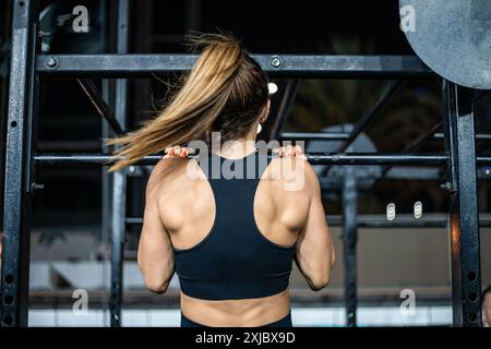 A woman in a black sports bra is doing pull-ups on a pull-up bar in a gym. Her blonde hair is pulled back in a ponytail. Stock Photo