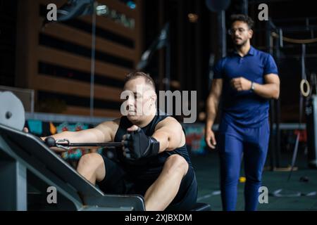 A man works out on a rowing machine at a gym. Another man stands behind him, likely an instructor, observing his form. Stock Photo